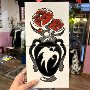 Op de afbeelding: Decoratieve tegel met een zwart-witte vaas met een gestileerd bloemmotief. De vaas bevat rode bloemen met grijze accenten, tegen een witte achtergrond. De tegel wordt door een hand vastgehouden.