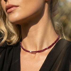 Puede incluir: Un delicado collar con pequeñas cuentas redondas de color rojo intenso. El collar se lleva en el cuello de una persona, y se ve un único y pequeño acento brillante. La persona lleva una blusa negra.