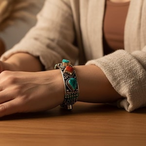 May include: A silver cuff bracelet featuring turquoise and coral-coloured stones. The bracelet has detailed metalwork and is shown on a wooden surface. The background includes neutral tones and decorative vases.