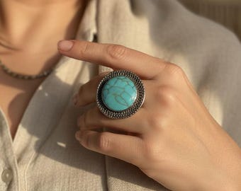 Puede incluir: Un anillo turquesa grande y redondo con un engaste plateado texturizado. El anillo presenta una vibrante piedra azul-verdosa. Esta joya es un accesorio llamativo.