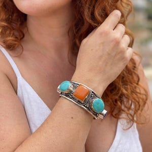 Puede incluir: Una pulsera de brazalete de plata con una piedra naranja cuadrada central flanqueada por dos piedras turquesas ovaladas. La banda de metal muestra intrincados detalles de alambre trenzado y cuentas que rodean cada una de las tres gemas engastadas.