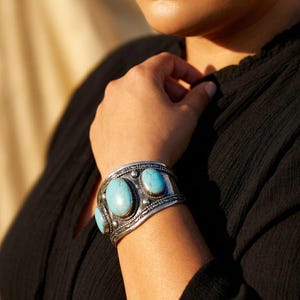May include: A silver cuff bracelet featuring three large, oval turquoise stones. The bracelet has a detailed, textured design around the stones and edges. The jewellery is worn on a person's wrist, with a black top in the background.