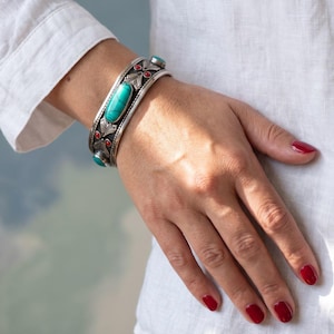 May include: A silver cuff bracelet featuring a large oval turquoise stone in the centre, flanked by smaller turquoise accents and inlaid with tiny red stones. The metal surface displays intricate engraved patterns and a darkened oxidised finish.