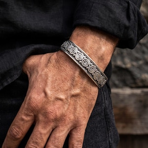 May include: A silver cuff bracelet featuring a repeating pattern of circular spiral motifs and small raised dots. The metal has a darkened, oxidised finish within the recessed areas of the design, highlighting the intricate texture of the metalwork.