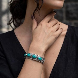 May include: A silver cuff bracelet featuring four oval-shaped turquoise stones set in a row. The metal band displays intricate decorative detailing along the edges and around each stone setting, creating a textured appearance on the wrist.