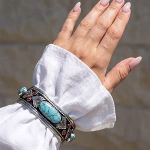 May include: A silver cuff bracelet featuring a large, oval, blue-green turquoise stone in the centre, flanked by smaller turquoise stones and accented with small, round, red coral inlays. The metal surface displays intricate engraved floral patterns.