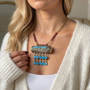 May include: A necklace featuring a central cylindrical metal pendant with turquoise-colored inlays and five dangling rows of matching circular turquoise-colored charms. The chain consists of small dark red beads and gold-toned metallic beads.