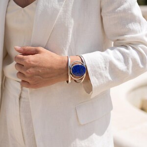 May include: A silver cuff bracelet featuring a large, oval-shaped, deep blue lapis lazuli stone set in a polished metal frame. The bracelet has a multi-layered, open-work design with curved silver bands that wrap around the wrist.