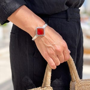 May include: A silver cuff bracelet featuring a square-shaped, deep red stone set in a decorative frame. The bracelet has a polished metallic finish and a textured border surrounding the central red gemstone, creating a distinct contrast.