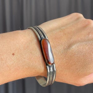 Pode incluir: Uma pulseira tipo bracelete de tom prateado com uma pedra oval central, lisa e alongada, de cor castanho-avermelhada escura. A banda de metal apresenta um design texturizado e estriado com três linhas paralelas ao longo do bracelete.