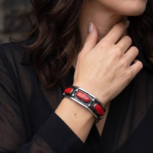 Può includere: Un bracciale rigido in argento con tre pietre rosse di forma ovale incastonate in metallo scuro. Il bracciale presenta una fascia in argento lucido con piccole sfere argentate posizionate tra ogni pietra, creando un design strutturato.