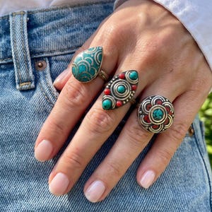 May include: Three silver rings featuring inlaid turquoise and red stone accents. One ring is teardrop-shaped with a scalloped pattern, one is an elongated oval with circular stone settings, and one is a round floral-inspired design with a central stone.