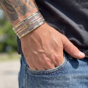 Pode incluir: Uma pulseira larga tipo bracelete com um design multimetálico, apresentando faixas alternadas de cobre, latão e metal prateado. A superfície é decorada com intrincados padrões de espinha de peixe gravados e texturas trançadas ao longo da peça.