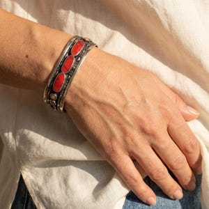 May include: A silver cuff bracelet featuring three prominent oval-shaped red stones set in a row, accented by smaller circular stones and intricate metal detailing along the band, creating a textured and layered appearance on the wrist.