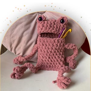 May include: A handmade, pink crocheted frog-shaped storage container. The frog has large, black eyes and holds crochet hooks in its open mouth. The container is sitting on a white surface with a pink pillow in the background.