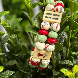 May include: A beaded bracelet featuring cream-colored rectangular beads with black markings and round beads in red, green, and cream with floral designs. The bracelet is set against a backdrop of green foliage.