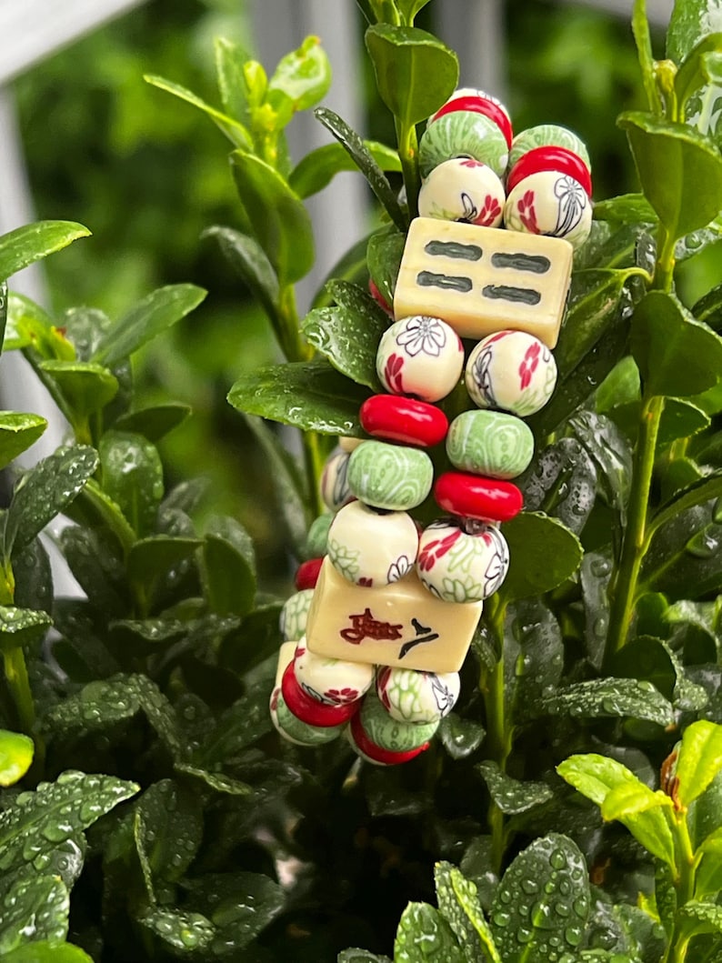 May include: A beaded bracelet featuring cream-colored rectangular beads with black markings and round beads in red, green, and cream with floral designs. The bracelet is set against a backdrop of green foliage.