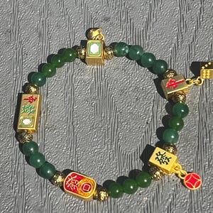 May include: A beaded bracelet featuring dark green round beads interspersed with gold-toned metal spacers and rectangular charms decorated with red, green, and black Chinese characters and symbols, including a small gold die and a red circular charm.