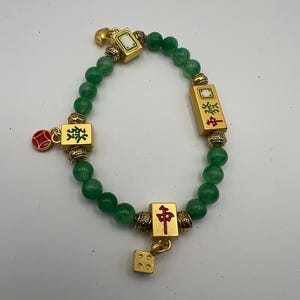 May include: Green beaded bracelet with gold-coloured accents. The bracelet features gold-coloured charms with Chinese characters and a small red charm. A gold-coloured dice charm hangs from the bracelet.