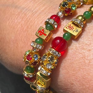 May include: Close-up of two gold-coloured beaded bracelets. The bracelets feature red, green, and clear crystal beads, along with gold-coloured square beads with red and gold symbols. The bracelets are on a person's wrist.