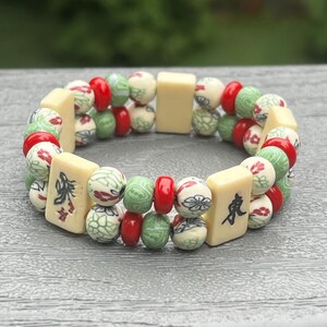 May include: A beaded bracelet featuring a mix of cream, red, and green beads. The bracelet includes rectangular cream-colored tiles with Chinese characters and round beads with floral and geometric patterns. The bracelet is on a gray surface.