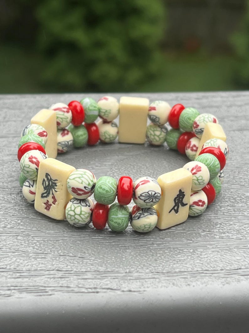 May include: A beaded bracelet featuring a mix of cream, red, and green beads. The bracelet includes rectangular cream-colored tiles with Chinese characters and round beads with floral and geometric patterns. The bracelet is on a gray surface.