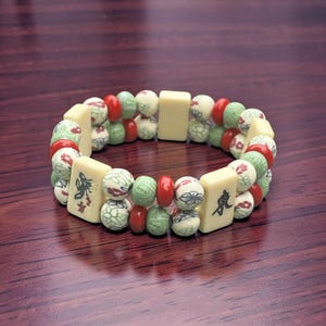 May include: A beaded bracelet featuring a mix of ivory-colored rectangular and round beads. The round beads are decorated with red, green, and white patterns. The bracelet has a dark wood background.