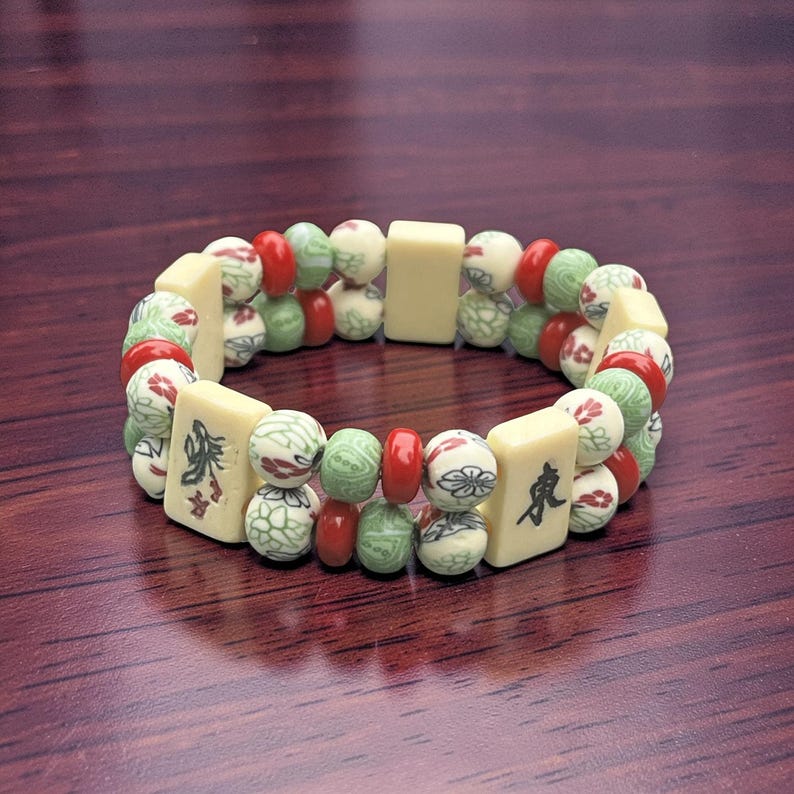 May include: A beaded bracelet featuring a mix of ivory-colored rectangular and round beads. The round beads are decorated with red, green, and white patterns. The bracelet has a dark wood background.