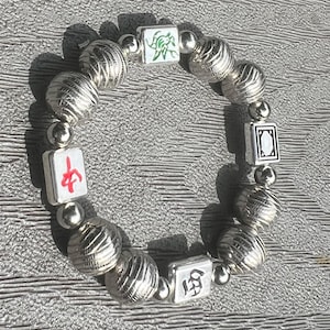 May include: A stretch bracelet featuring alternating silver-toned ribbed spherical beads and small smooth round beads, interspersed with four square white tiles. The tiles display a green bamboo design, a red Chinese character, a black symbol, and a black border.