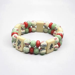 May include: A double-strand bracelet featuring cream-colored rectangular tiles with black characters, interspersed with red, green, and cream-colored round beads. The beads have floral and geometric patterns.