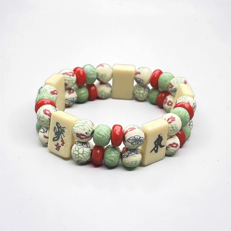 May include: A double-strand bracelet featuring cream-colored rectangular tiles with black characters, interspersed with red, green, and cream-colored round beads. The beads have floral and geometric patterns.