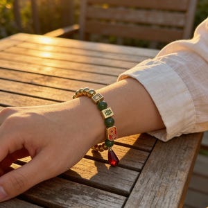 May include: A gold and green beaded bracelet with red accents. The bracelet features gold beads, green square beads with symbols, and a red teardrop-shaped charm. The bracelet is worn on a wrist, with a wooden table in the background.