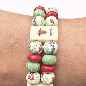 May include: A beaded bracelet featuring alternating red, green, and cream-colored beads. The cream beads are decorated with red floral patterns and black accents. Two rectangular tiles with red symbols and lines are also included.