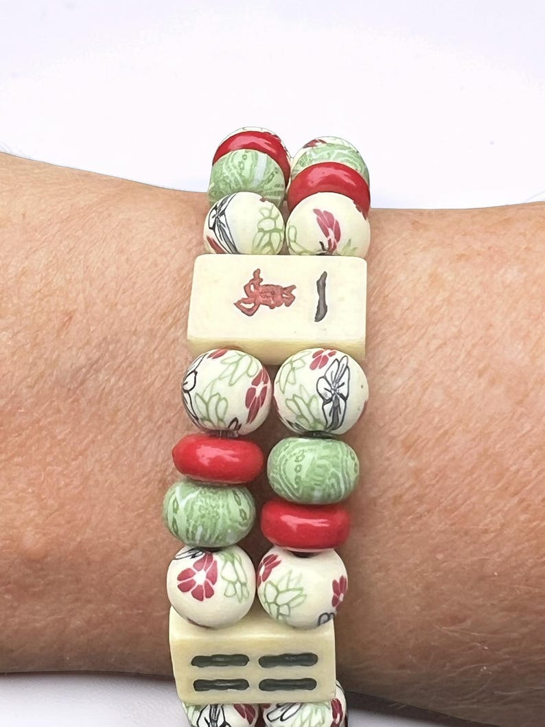 May include: A beaded bracelet featuring alternating red, green, and cream-colored beads. The cream beads are decorated with red floral patterns and black accents. Two rectangular tiles with red symbols and lines are also included.