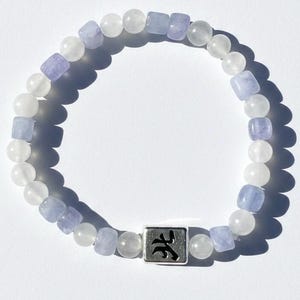Op de afbeelding: Een kralenarmband met afwisselend ronde, witte, doorschijnende kralen en vierkante, lichtpaarse kralen. Een vierkante, zilverkleurige metalen kraal is onderaan gecentreerd en bevat een zwart gegraveerd teken dat lijkt op het kanji voor water.