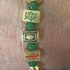 May include: A gold bracelet with green beads and rectangular charms. The charms feature red and green designs, including Chinese characters. A red, teardrop-shaped charm hangs from the bracelet.