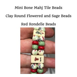 May include: A close-up of a bracelet featuring mini bone Mahj tile beads, clay round flowered and sage beads, and red rondelle beads. The bracelet has a mix of cream, red, and green beads. The text "一萬" is visible on one of the bone tile beads.
