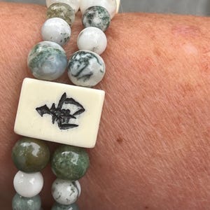 May include: A double-strand bracelet featuring round beads in shades of white, moss green, and grey with marbled patterns. The bracelet includes a rectangular off-white tile bead engraved with a black Chinese character, creating a layered design.