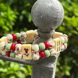 May include: A beaded bracelet featuring a mix of red, green, and cream-colored beads. The bracelet includes rectangular blocks with Chinese characters and floral patterns. The background is a blurred green.