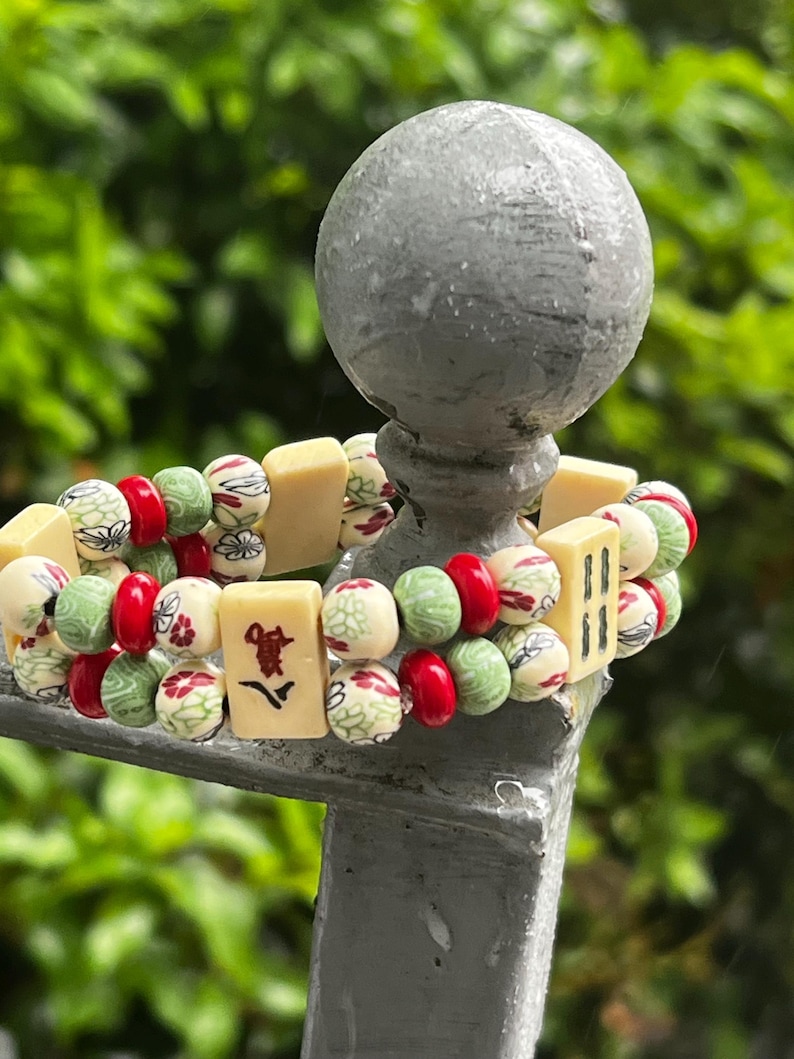 May include: A beaded bracelet featuring a mix of red, green, and cream-colored beads. The bracelet includes rectangular blocks with Chinese characters and floral patterns. The background is a blurred green.