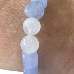 Op de afbeelding: Een elastische armband met afwisselend gladde, ronde en getrommelde kralen in tinten lichtblauw en doorschijnend wit. De armband bevat een enkele metallic zilverkleurige bolvormige kraal en een rechthoekig wit accentstuk.