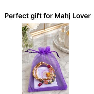 May include: A gold beaded bracelet with red and black accents, displayed inside a purple mesh gift bag. The bracelet features decorative charms. Text on the image reads "Perfect gift for Mahj Lover".