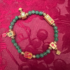 May include: A jade green beaded bracelet with gold-coloured accents and charms. The charms include rectangular and square pieces with red and white details, and a small gold-coloured die. The bracelet is on a red background.