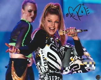 May include: A woman with blonde hair in a high ponytail, wearing a black and white patterned outfit, holds a gold microphone. She is smiling and wearing multiple rings. A second person is visible in the background.