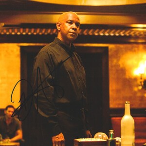 May include: A bald person wearing a black long-sleeved button-down shirt stands in a dimly lit room with warm golden lighting. A black ink autograph is visible across the center of the image. A clear glass bottle sits on a surface in the foreground.