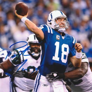May include: A football player in a blue and white uniform throws a brown football. The jersey has the number 18 in white. Other players in white and green uniforms are in the background. The image is signed in blue ink.