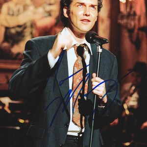 May include: A photograph of a person with curly dark hair wearing a dark charcoal suit jacket, a crisp white collared shirt, and a patterned orange and brown necktie. The person is holding a black microphone on a stand, with a blue ink signature.