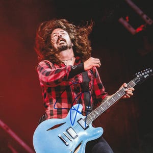 May include: A musician with long brown hair in motion, wearing a red and black plaid button-down shirt, playing a light blue semi-hollow body electric guitar with a Gibson headstock logo. A blue ink signature is visible on the guitar body.