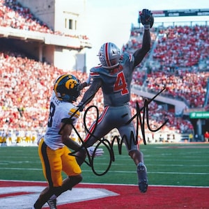 May include: A football player in a gray uniform with the number 4 and a helmet with American flag decals jumps to catch a ball, while a player in a white jersey with the number 8 and yellow pants defends on a green field.