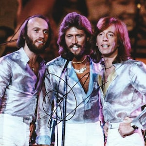 May include: A photograph of three individuals with long hair and beards wearing shimmering silver long-sleeved shirts and white trousers. The individuals are positioned side-by-side, with a black microphone stand in front of the center figure.